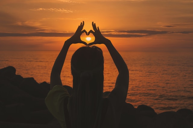 Woman faced to sunset and making a heart with her fingers around the Sun