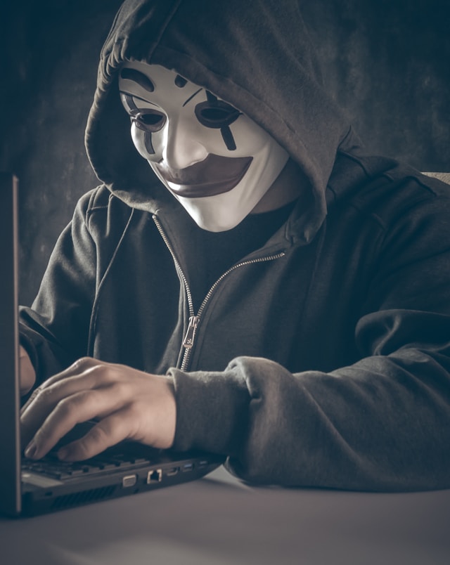 Photo of a criminal looking person, wearing a mask, while sitting behind a laptop